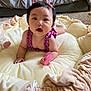 baby, child, infant, purple_clothing, pink_bows, soft_cushion, carpet, bed, indoor, cute, holding_object, pink_object, portrait, face, hands, wide_eyes, open_mouth, skin, short_hair, person