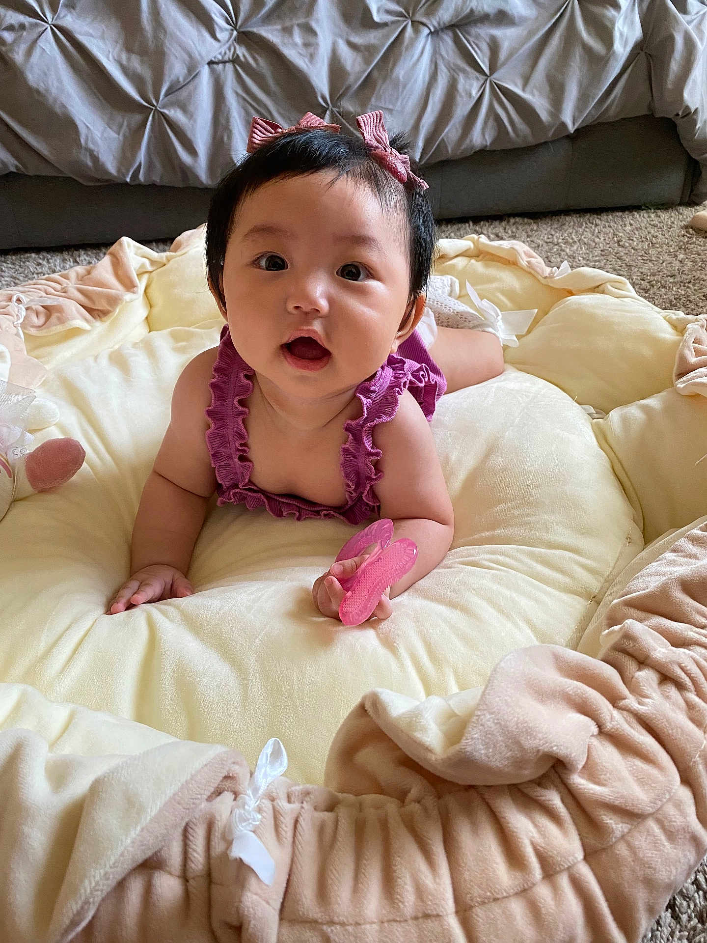 Selene is registered to the contest to win money with this photo: baby, child, infant, purple_clothing, pink_bows, soft_cushion, carpet, bed, indoor, cute, holding_object, pink_object, portrait, face, hands, wide_eyes, open_mouth, skin, short_hair, person