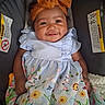 baby, child, smiling, orange_bow, floral_dress, car_seat, warning_label, cute, portrait, infant, happy, curly_hair, clothing, seatbelt, indoor, person, young_child, headband, baby_girl, colorful