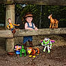 action_figures, boots, bullseye, buzz_lightyear, child, cowboy_hat, denim_overalls, gravel, mr_potato_head, outdoor, portrait, rex, slinky_dog, smiling, toddler, toy_horse, toy_story, wooden_fence, woody, zoo_like_setting
