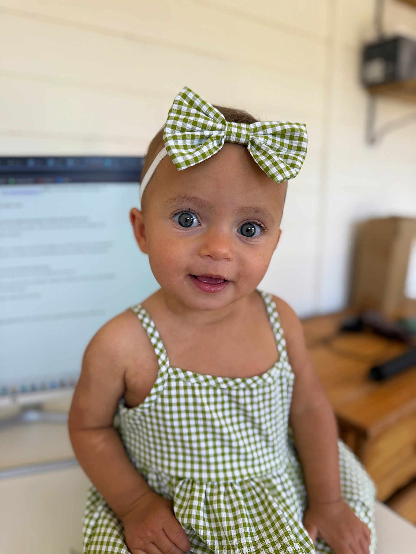 Etta is registered to the contest to win money with this photo: baby, child, girl, blue_eyes, bow, headband, green_checkered, dress, sitting, indoor, computer_screen, blurred_background, face, smile, person, cute, portrait, infant, skin, hand