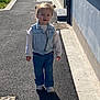 child, toddler, denim_vest, jeans, white_sneakers, outdoor, sidewalk, sunny, shadow, hair_buns, walking, casual_clothing, smiling, pavement, daylight, urban, person, small_child, cute, happy