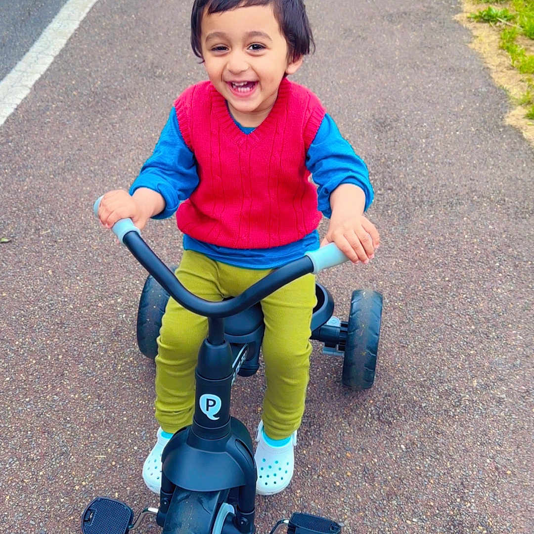 Arshman is registered to the contest to win money with this photo: casual_clothing, child, fun, green_pants, happy, joy, outdoor, park, pathway, pavement, person, play, recreation, red_vest, smiling, toddler, toy, tricycle, white_shoes, young_child