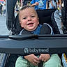 baby, child, stroller, smile, happy, outdoor, people, clothing, shoe, hand, street, daylight, person, blue_shirt, green_pants, bottle, background, urban, parked_stroller, babytrend