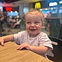 Manaia is registered to the contest to win money with this photo: toddler, child, smiling, happy, curly_hair, table, wooden_table, indoor, restaurant, fast_food, mcdonalds_sign, blurry_background, seated, short_sleeve_shirt, light_pink_shirt, face, person, baby, cute, joyful