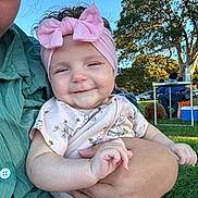 Manaia joined the competition — help win amazing prizes! baby, child, person, pink_bow, headband, smile, holding, adult_hand, outdoor, grass, tree, park, daylight, shirt, cute, happy, nature, people, relaxation, family