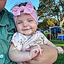 baby, child, person, pink_bow, headband, smile, holding, adult_hand, outdoor, grass, tree, park, daylight, shirt, cute, happy, nature, people, relaxation, family
