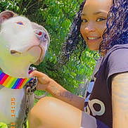 Lily is registered to the contest to win money with this photo: casual_clothing, crouching, curly_hair, daylight, dog, friendship, grass, greenery, happy, leash, nature, outdoor, pet, portrait, rainbow_collar, smile, summer, sunlight, tattoo, woman