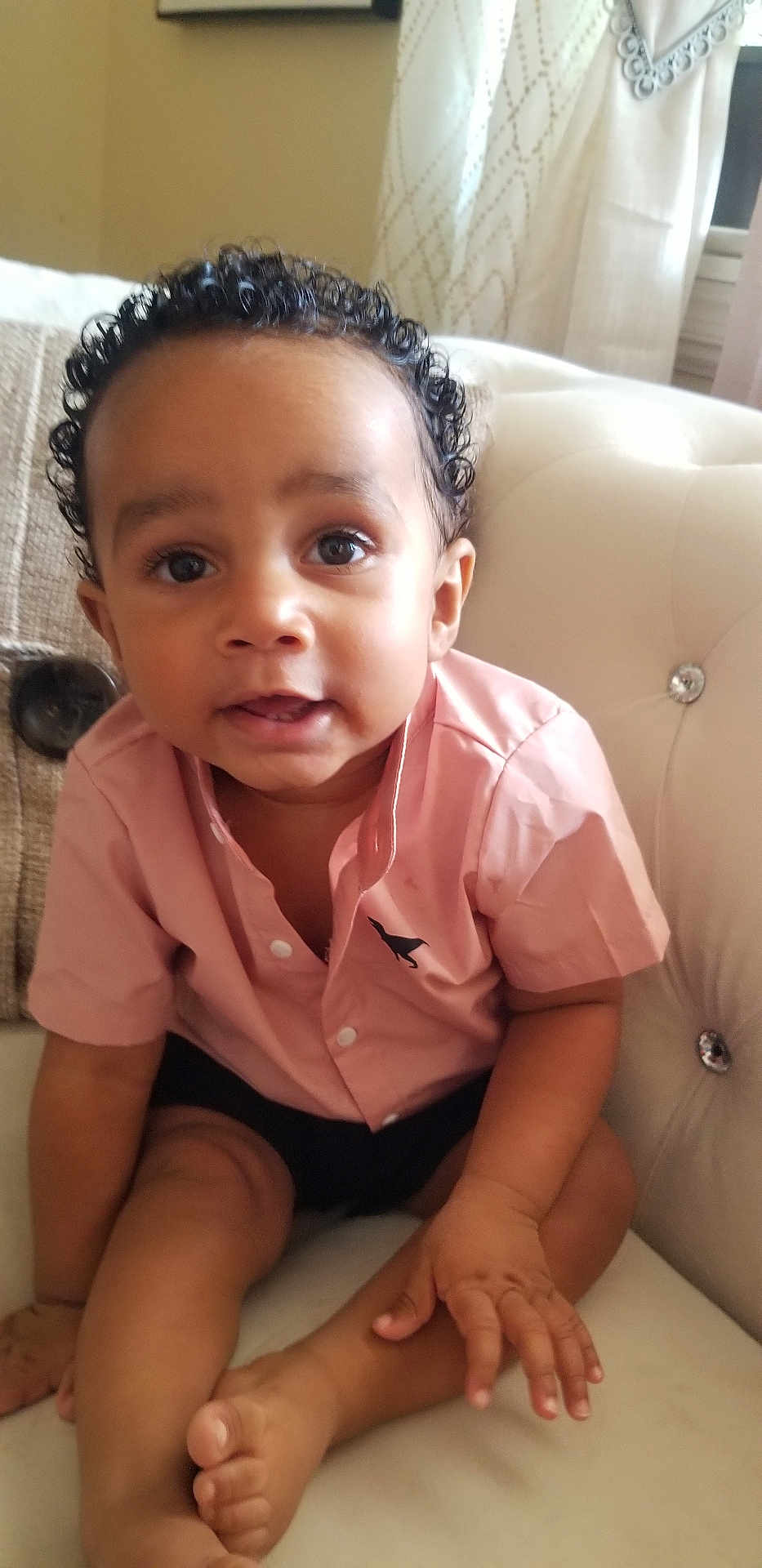 Jeremias is registered to the contest to win money with this photo: toddler, child, curly_hair, pink_shirt, black_shorts, couch, indoor, sitting, hand, foot, face, eyes, skin, person, baby, cute, expression, comfort, home, furniture
