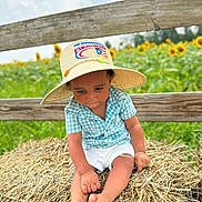 Jeremias is registered to the contest to win money with this photo: toddler, child, hat, straw_hat, blue_shirt, shorts, barefoot, hay, hay_bale, fence, sunflower, flower_field, outdoor, grass, summer, nature, portrait, sitting, sunny, rural