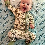 Ashton is registered to the contest to win money with this photo: baby, smiling, onesie, infant, bed, sheet, pattern, text, happy, child, footed_pajamas, face, playful, cute, lying_down, person, indoors, apparel, fabric, pajamas