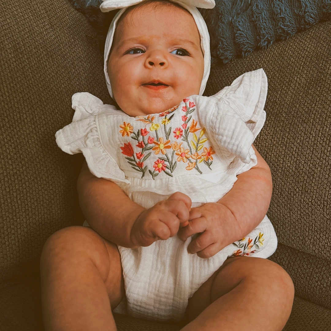 Wren is registered to the contest to win money with this photo: baby, infant, child, couch, headband, white_outfit, floral_embroidery, cute, smiling, sitting, indoor, plush_pillow, cozy, portrait, small_hands, legs, comfortable, soft_texture, happy, adorable