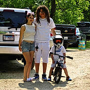 Josiah is registered to the contest to win money with this photo: adult, bicycle, casual_clothing, child, daytime, dirt_road, family, glasses, greenery, helmet, helmet_visor, outdoor, parked_cars, posing, shorts, sneakers, summer, sunlight, trees, tshirt