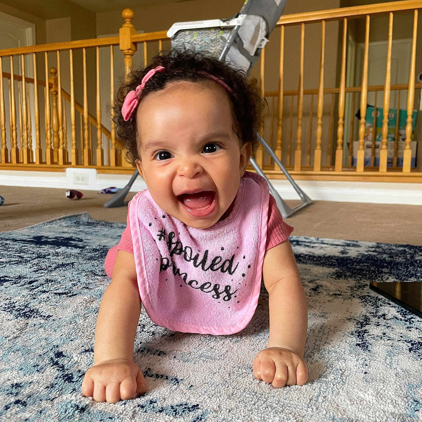 Jenysis is registered to the contest to win money with this photo: baby, baby_chair, carpet, child, clothing, crawling, cute, expression, floor, happy, headband, home, indoor, infant, person, pink_bib, playful, rug, smiling, wooden_railings