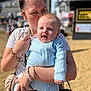 baby, woman, child, holding, kiss, outdoor, beach, sunlight, blue_clothing, happy, person, portrait, sand, daytime, casual_wear, accessory, bracelet, handbag, blurred_background, smile