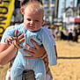 John is registered to the contest to win money with this photo: baby, child, person, holding, blue_clothing, knitwear, outdoor, sunny, sand, blurred_background, building, adult_hands, portrait, cute, young_child, daylight, casual, human, face, infant