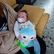 François participe au concours pour gagner de l'argent avec cette photo : baby, pacifier, plush_toy, couch, adult, clothing, indoor, floor, person, holding, toy, child, seated, comfort, young, casual_wear, blue_pants, beige_jacket, relaxation, home