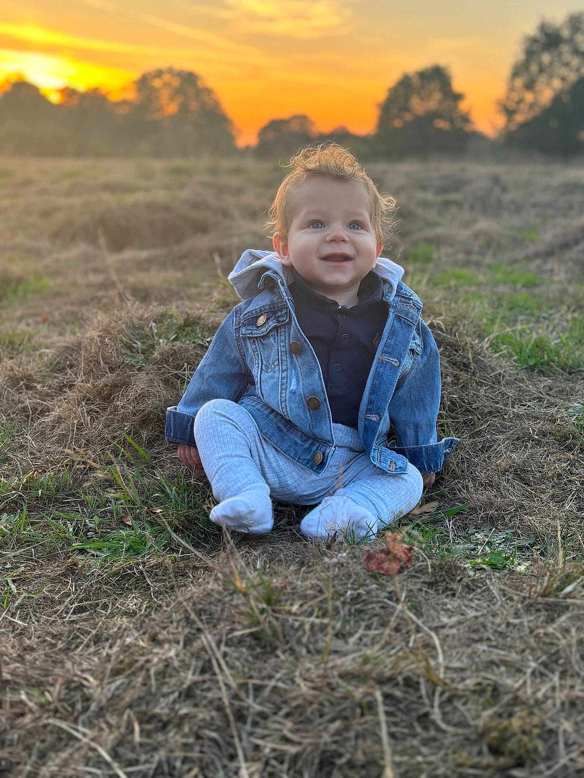 Otto is registered to the contest to win money with this photo: baby, child, denim_jacket, grass, sunset, outdoor, smiling, curly_hair, nature, field, clothing, happy, sitting, person, portrait, casual_wear, blue_eyes, daylight, young_child, scenery
