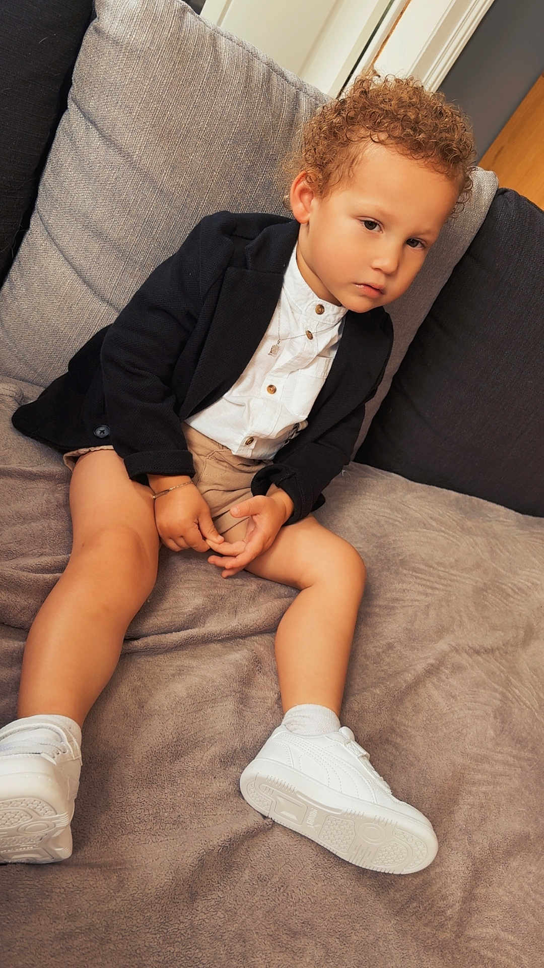 Cameron participe au concours pour gagner de l'argent avec cette photo : child, kid, boy, curly_hair, blazer, white_shirt, shorts, white_sneakers, socks, couch, blanket, indoor, portrait, seated, casual, fashion, accessory, bracelet, necklace, thoughtful