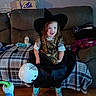 baby_bottle, blanket, child, couch, girl, halloween, living_room, missing_tooth, parquet_floor, plaid_blanket, playful_expression, pumpkin_bucket, red_hair, remote_control, smiling, taco_bell_box, teal_socks, toddler_shoes, witch_costume, witch_hat