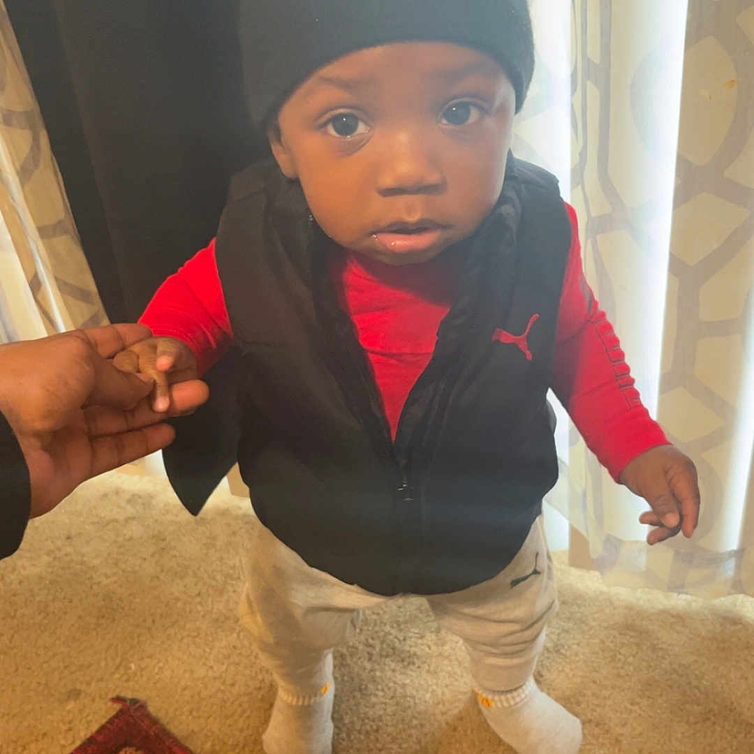 Jamir joined the competition — help win amazing prizes! toddler, child, indoor, carpet, curtain, beanie, vest, socks, holding_hand, person, standing, red_shirt, pants, adult_hand, floor, patterned_curtain, looking_up, curious, home, casual_clothing
