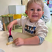 Sandro participe au concours pour gagner de l'argent avec cette photo : child, toddler, smiling, cookie, icing, brush, table, indoor, clothing, person, happy, playroom, background, furniture, yellow_chair, food, activity, cute, young_child, fun