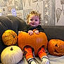 baby, pumpkin, halloween, child, orange_pants, socks, couch, blanket, smile, curly_hair, carved_pumpkin, white_pumpkin, decor, wall_art, indoor, happy, festive, cute, holiday, sitting