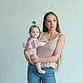 woman, baby, child, holding, portrait, indoor, casual_clothing, jeans, pink_top, long_hair, pony_tail, barefoot, light_background, minimalistic, mother_and_child, curious_expression, blue_eyes, human, family, gentle_lighting