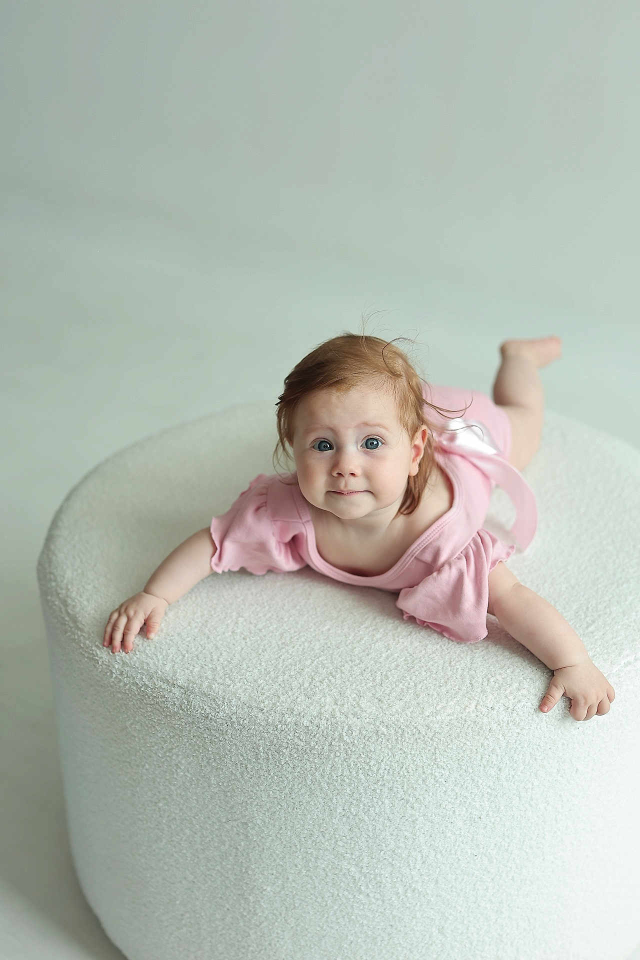 Emiliya joined the competition — help win amazing prizes! baby, infant, pink_dress, blue_eyes, curly_hair, ottoman, white, soft_texture, child, portrait, cute, indoor, furniture, smiling, expression, person, young_child, hands, feet, light_background