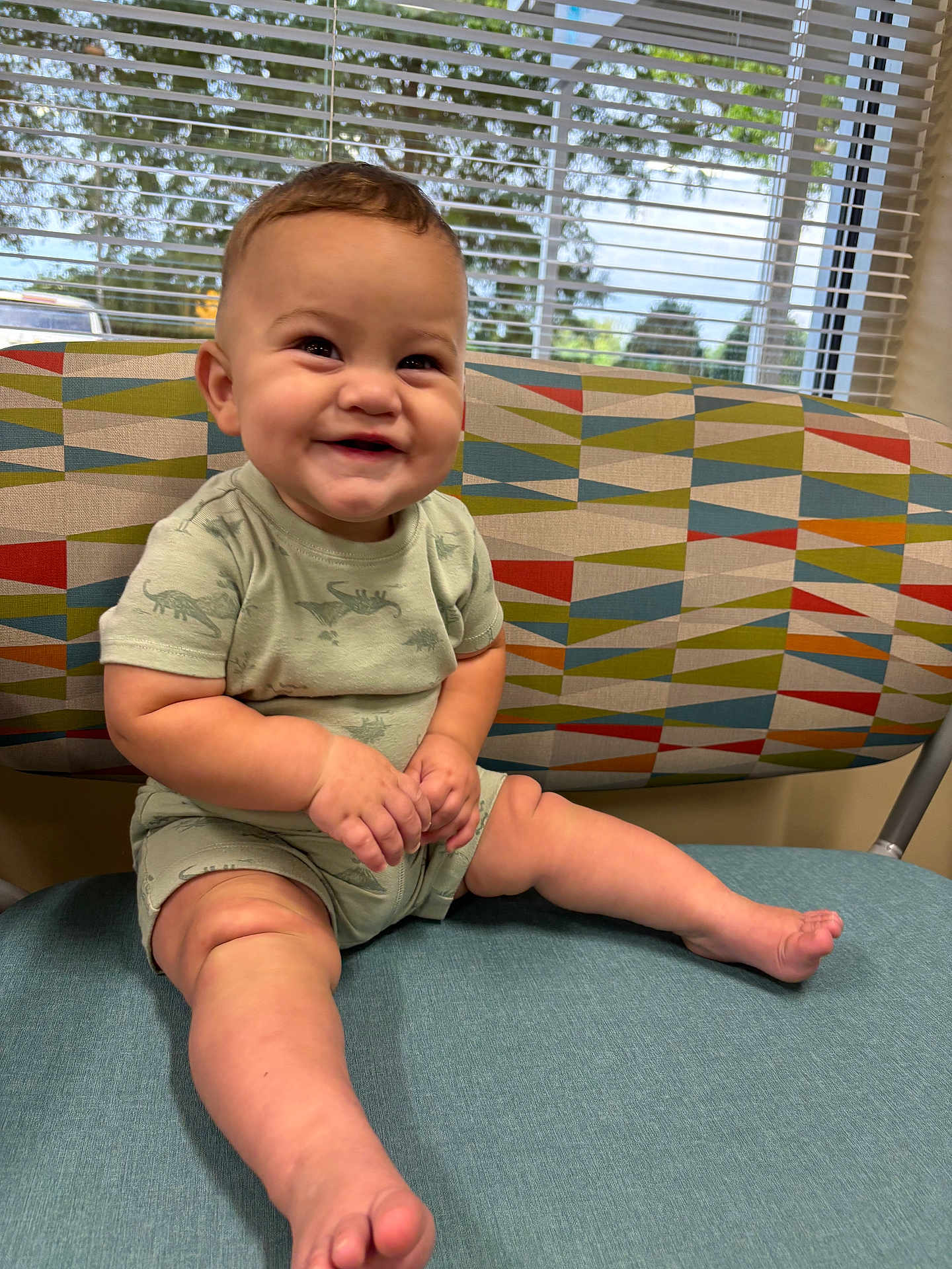 Ezekiel is registered to the contest to win money with this photo: baby, child, smiling, sitting, indoor, chair, patterned_fabric, window, blinds, natural_light, cute, happy, infant, legs, feet, shorts, tshirt, face, skin, person