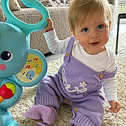 Judith a rejoint le concours — aidez-le/la à gagner de superbes lots ! baby, child, blue_eyes, blonde_hair, knitwear, purple_clothing, toy, musical_toy, carpet, living_room, couch, blanket, indoor, smiling, cute, sitting, footwear, person, home, playtime