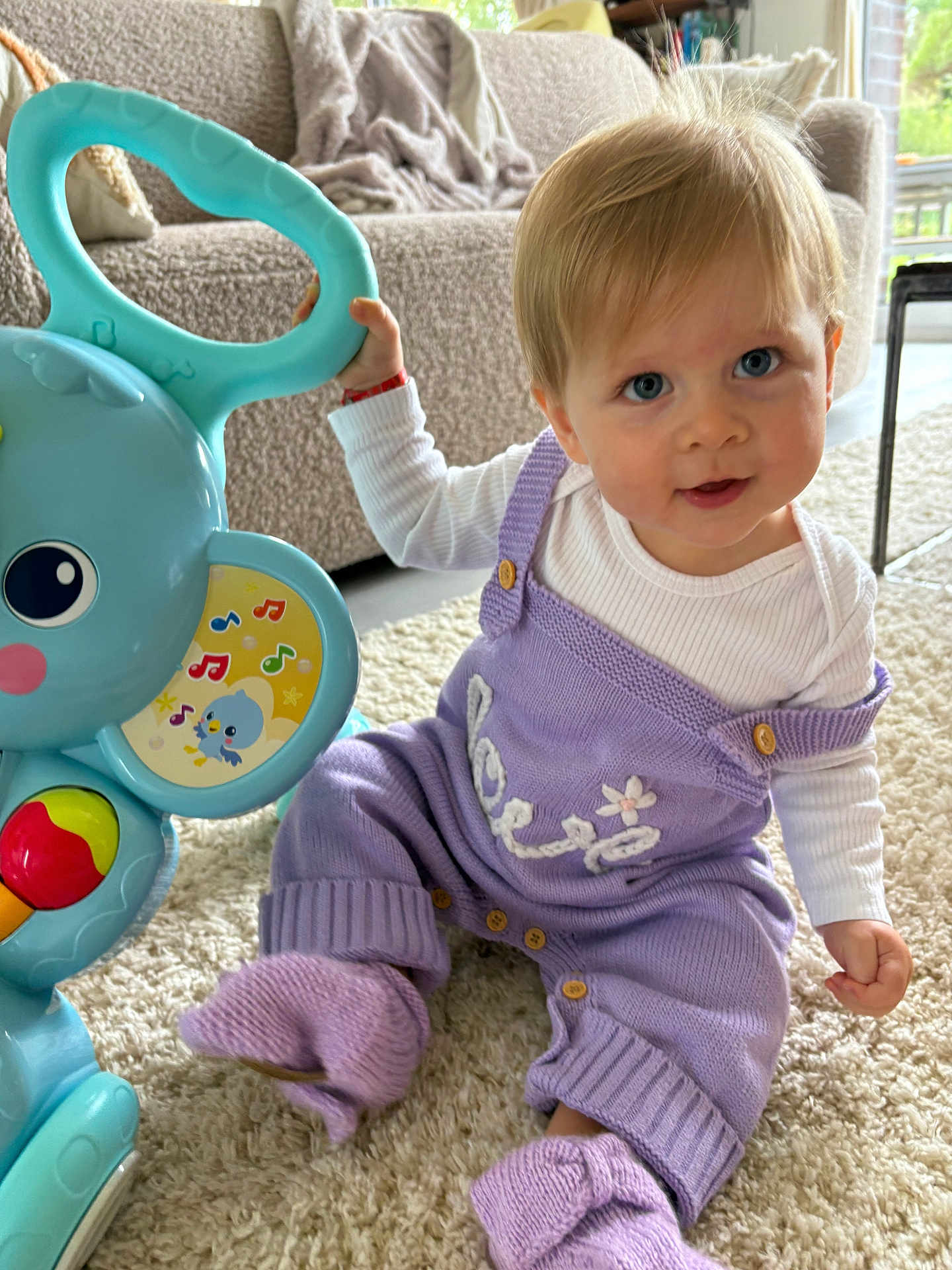 Judith a rejoint le concours — aidez-le/la à gagner de superbes lots ! baby, child, blue_eyes, blonde_hair, knitwear, purple_clothing, toy, musical_toy, carpet, living_room, couch, blanket, indoor, smiling, cute, sitting, footwear, person, home, playtime