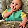 baby, high_chair, food, messy, bib, yellow_clothing, face, fingers, table, eating, crumbs, tray, indoor, child, curious, infant, mess, sitting, feeding, cute