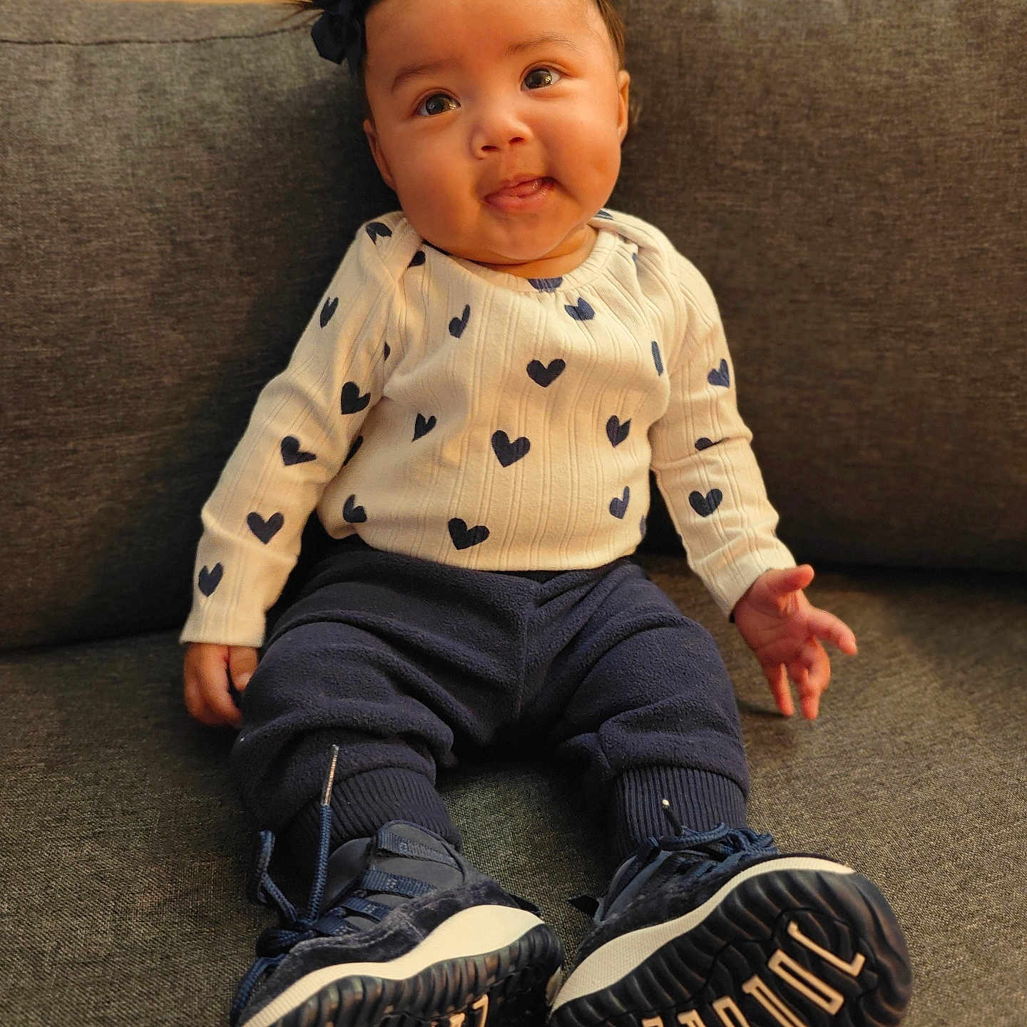 Navaya joined the competition — help win amazing prizes! baby, bows, child, cozy, cute, footwear, gray_couch, happy, hearts_pattern, indoor, navy_pants, pigtails, playful_expression, portrait, seated, smiling, sneakers, warm_lighting, white_sweater, young_child