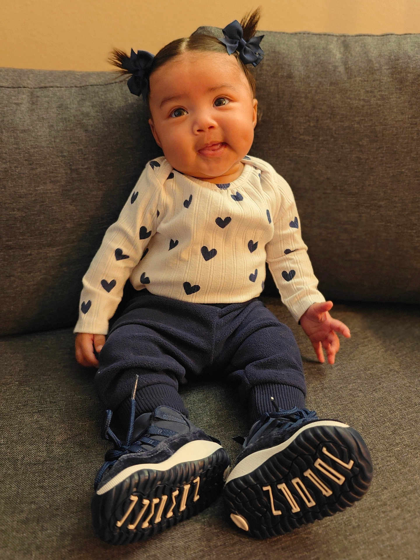 Navaya joined the competition — help win amazing prizes! baby, child, pigtails, bows, white_sweater, hearts_pattern, navy_pants, sneakers, gray_couch, indoor, cute, smiling, portrait, seated, playful_expression, young_child, footwear, cozy, warm_lighting, happy