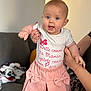 baby, child, person, pink_skirt, white_shirt, blue_eyes, hands, couch, blanket, indoor, smiling, cute, clothing, home, sitting, face, skin, furniture, holding, expression