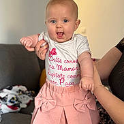 Bryana a rejoint le concours — aidez-le/la à gagner de superbes lots ! baby, child, smiling, pink_skirt, white_shirt, red_text, person, hand, couch, living_room, happy, indoor, clothing, cute, holding, adult_hand, face, portrait, infant, young_child