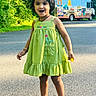 child, girl, green_dress, flower, smile, outdoor, sunlight, road, trees, food_truck, shoes, happy, portrait, nature, daytime, cute, standing, person, playful, casual