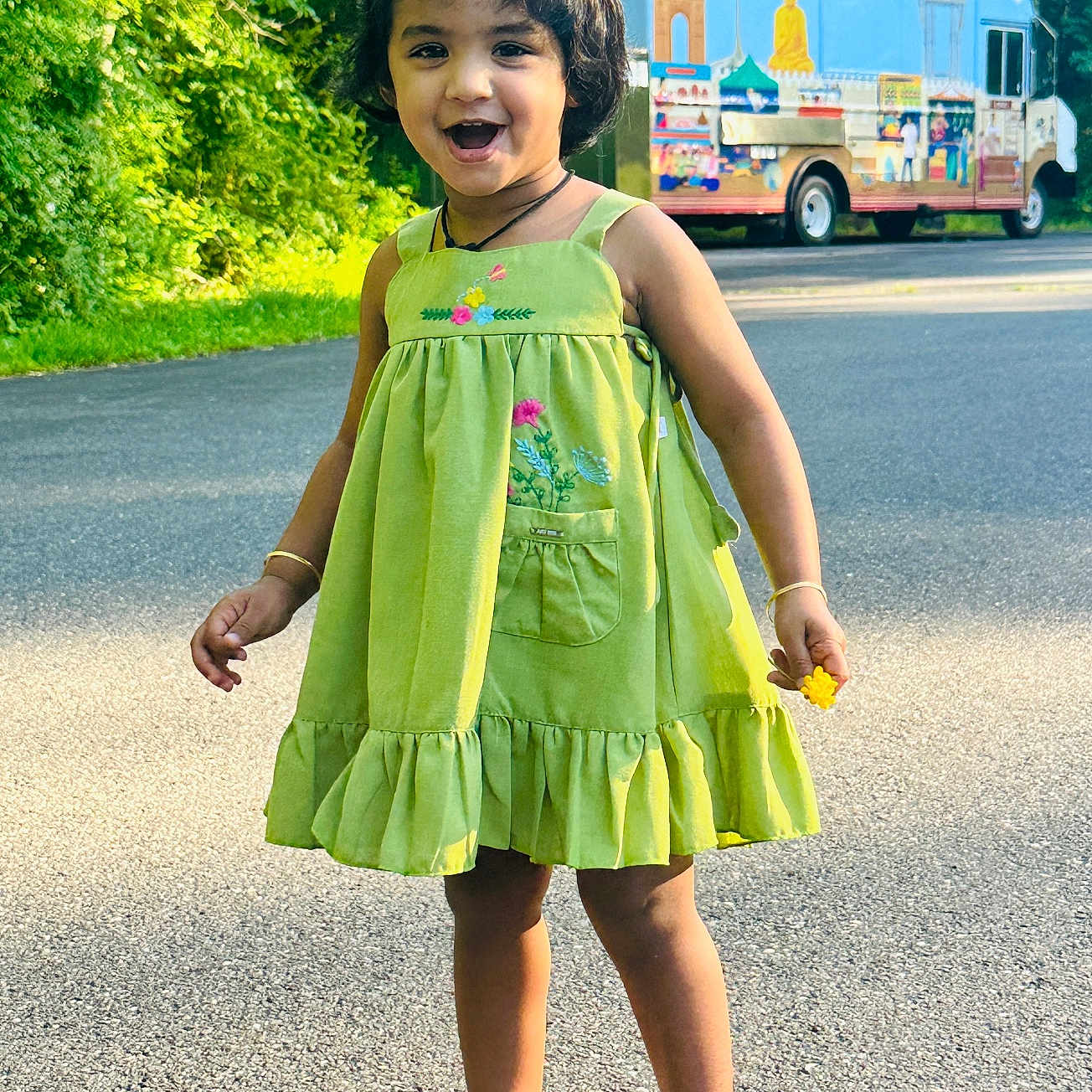 Nishika Sai joined the competition — help win amazing prizes! child, girl, green_dress, flower, smile, outdoor, sunlight, road, trees, food_truck, shoes, happy, portrait, nature, daytime, cute, standing, person, playful, casual
