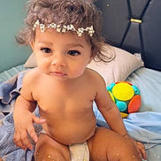 Elizabeth is registered to the contest to win money with this photo: baby, toddler, curly_hair, flower_headband, diaper, bed, blanket, toy, colorful, indoor, child, cute, skin, face, sitting, soft, sparkles, playful, head, person