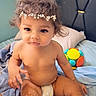 baby, toddler, curly_hair, flower_headband, diaper, bed, blanket, toy, colorful, indoor, child, cute, skin, face, sitting, soft, sparkles, playful, head, person