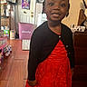 Nyla is registered to the contest to win money with this photo: child, girl, smile, red_dress, black_cardigan, indoor, wooden_floor, decor, bags, laundry_basket, wall_art, portrait, furniture, happy, home, person, hair_accessory, cabinet, floor_mat, cozy