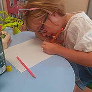 Jade a rejoint le concours — aidez-le/la à gagner de superbes lots ! art_supplies, blue_table, child, colorful, creative, drawing, focused, furniture, glasses, headband, indoor, paper, pencil, person, pink, table, toy, white_clothing, writing, young