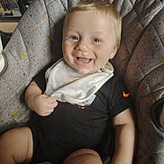 Kane is registered to the contest to win money with this photo: baby, smiling, bib, blond_hair, padded_seat, bracelet, happy, child, infant, clothing, face, person, indoor, cute, sitting, joyful, messy, comfort, beads, closeup