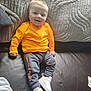 Kane joined the competition — help win amazing prizes! baby, toddler, child, smiling, sitting, clothing, orange_jacket, grey_pants, white_sneakers, under_armour, couch, leather, blanket, indoor, person, happy, footwear, fabric, young_child, portrait