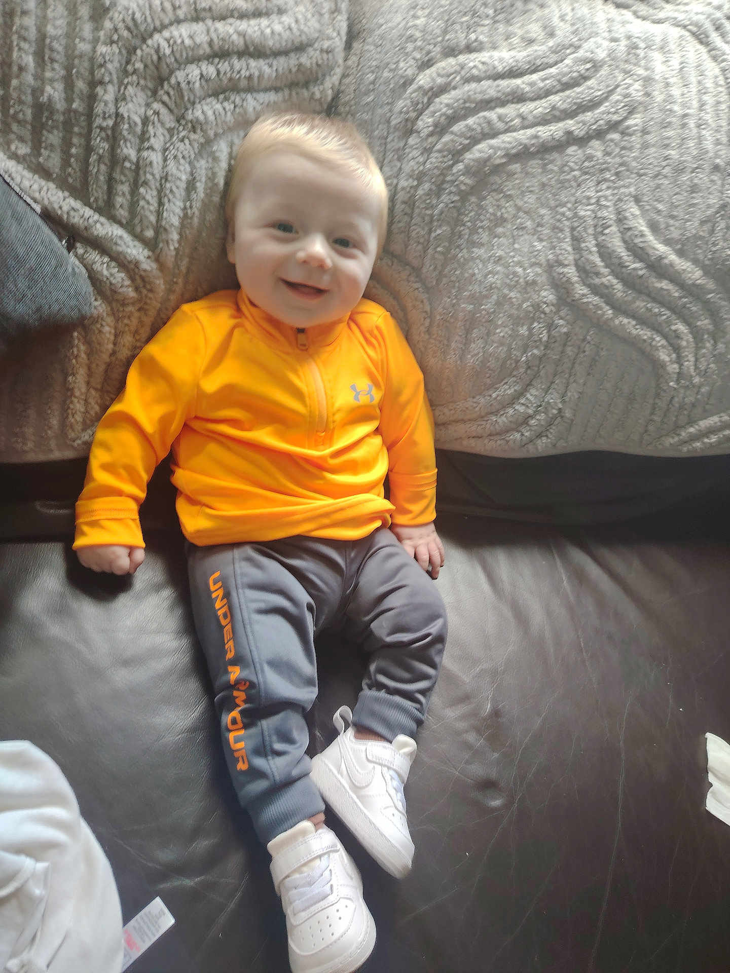Kane joined the competition — help win amazing prizes! baby, toddler, child, smiling, sitting, clothing, orange_jacket, grey_pants, white_sneakers, under_armour, couch, leather, blanket, indoor, person, happy, footwear, fabric, young_child, portrait