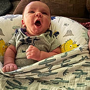 Jaxson is registered to the contest to win money with this photo: baby, infant, blanket, cactus_pattern, dinosaur_pillow, yawning, clothing, indoor, couch, cute, face, hand, person, newborn, soft_fabric, comfort, resting, expression, home, wrapped