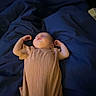 baby, infant, sleeping, lying_down, bed, blanket, blue_bedding, onesie, ribbed_clothing, arms_raised, face, portrait, indoors, bedroom, dim_lighting, cozy, peaceful, cute, newborn, photography