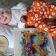 Ryleiah is registered to the contest to win money with this photo: baby, bejeweled_letter, child, closeup, colorful_art, cute, floral_pattern, flower, happy, headband, indoors, infant, lying_down, orange_pants, pillow, portrait, smiling, two_months, white_top, wooden_plaque
