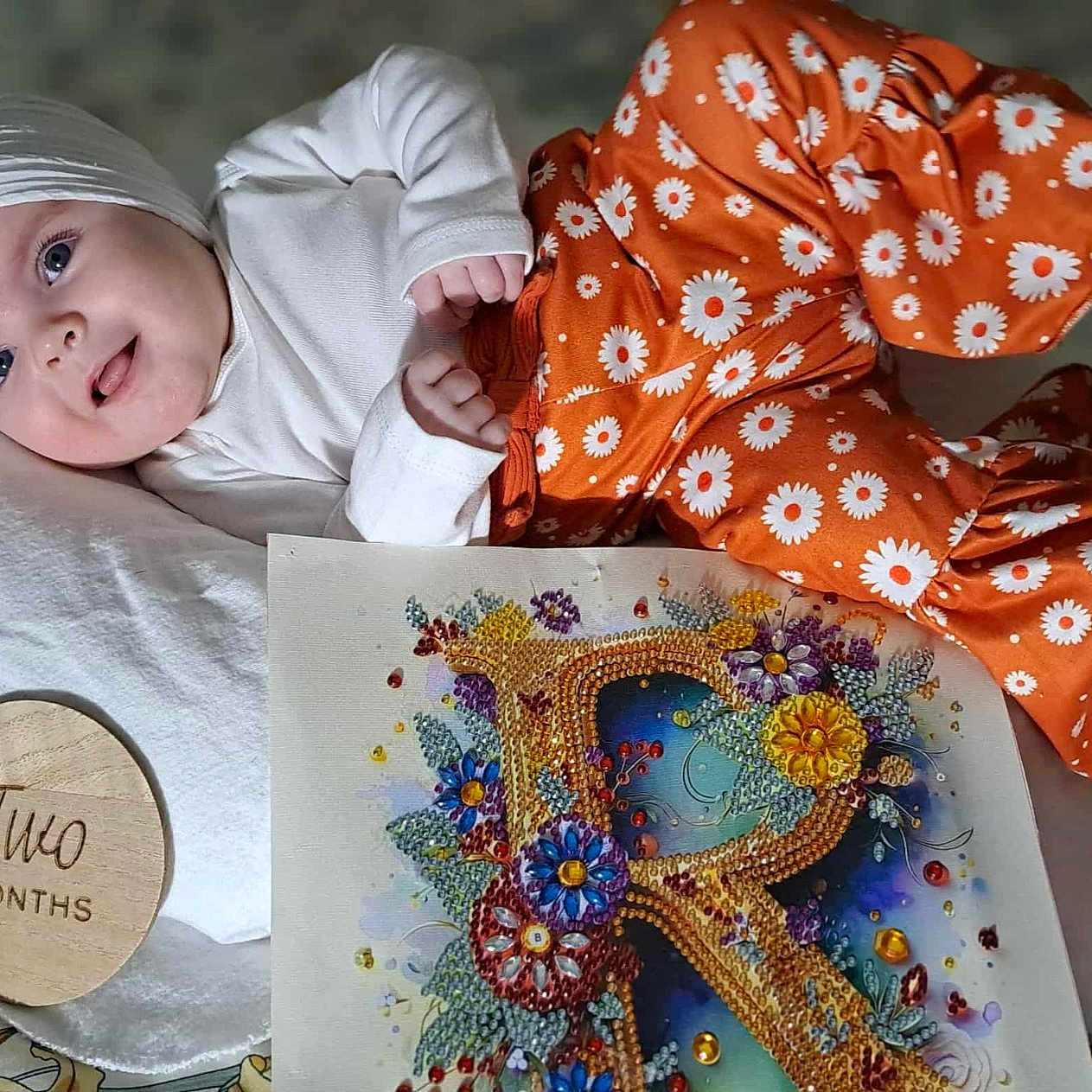 Ryleiah is registered to the contest to win money with this photo: baby, bejeweled_letter, child, closeup, colorful_art, cute, floral_pattern, flower, happy, headband, indoors, infant, lying_down, orange_pants, pillow, portrait, smiling, two_months, white_top, wooden_plaque